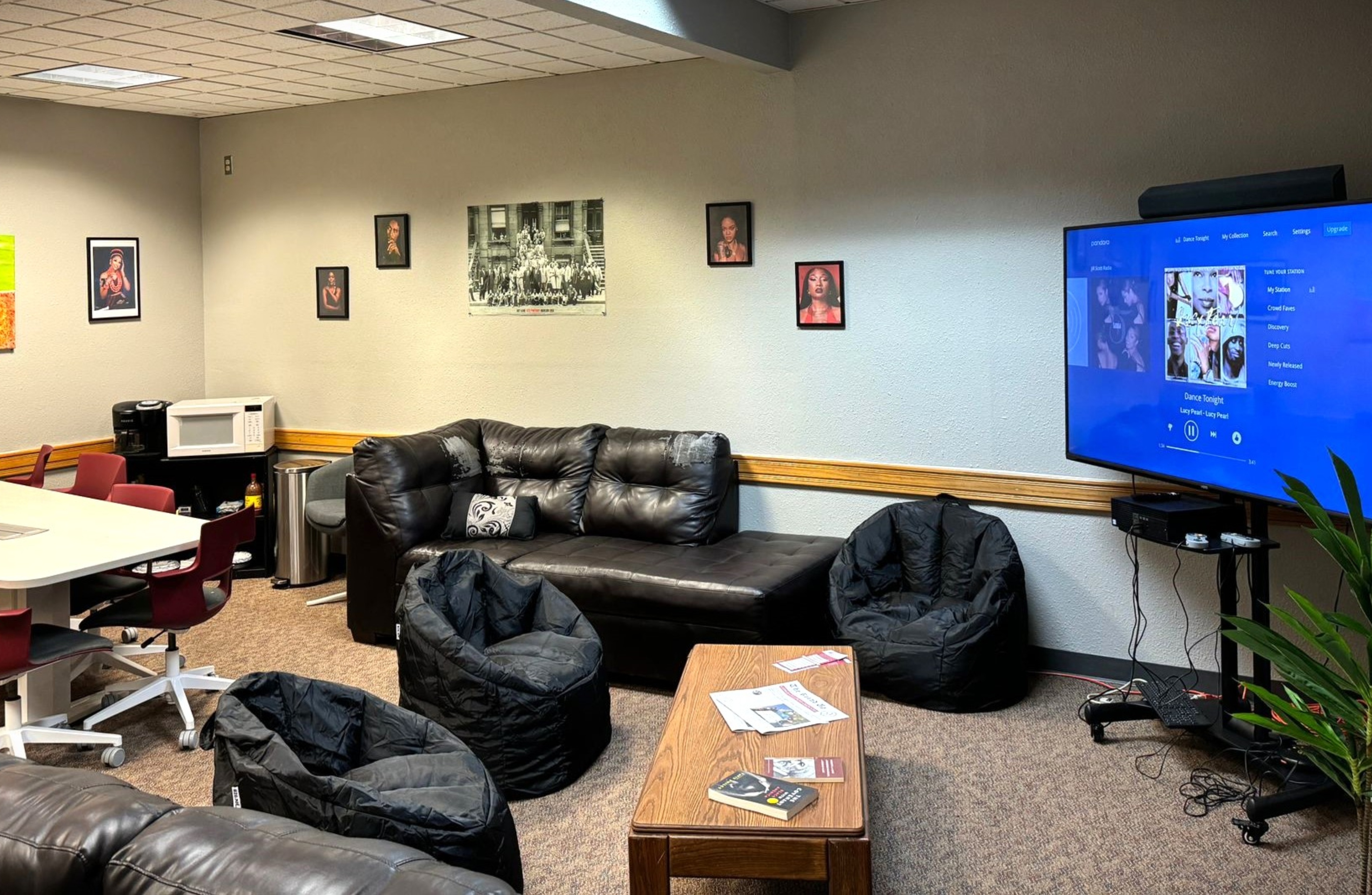 The image shows a cozy, multi-purpose room that combines a lounge and a small kitchenette. Prominent in the foreground is a black leather sectional sofa with a black and white throw pillow. Adjacent to the sofa are three black bean bag chairs arranged on a beige carpeted floor. In front of the seating area is a wooden coffee table with various items, including a few magazines and a book, scattered on it.  On the left side of the room, there is a white square table with four red swivel chairs surrounding it. Behind the table is a small kitchenette area with a white microwave on a black stand, along with other appliances and storage containers.  On the right side of the room, a large flatscreen TV is mounted on the wall. The TV is displaying a blue screen with music-related content, including various album covers and a list of songs. Below the TV is a small black shelf with electronic devices and a plant partially visible on the right edge of the frame.  The walls are decorated with several framed pictures, including one black and white historical photograph and multiple colored portraits. The ceiling is fitted with white square tiles and recessed lighting.