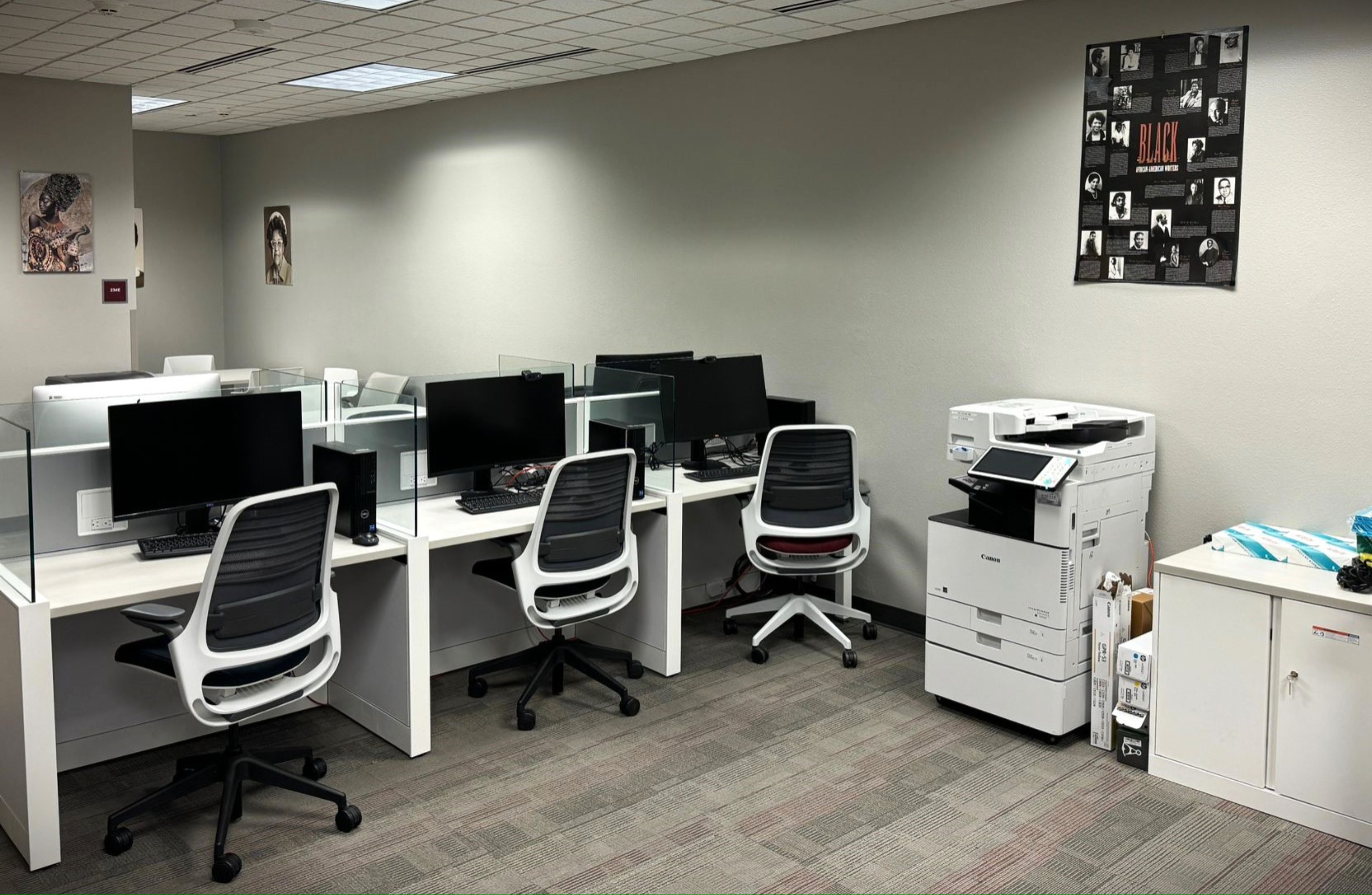 The image portrays a small, clean office space with a row of four workstations and a multifunction printer/copier. Each workstation consists of a white desk with a glass partition, a black computer monitor, a keyboard, and a white office chair with black cushions. To the right, there is a Canon office printer/copier next to a white cabinet. The floor has a grey and beige patterned carpet. The room is well-lit with recessed ceiling lights. The beige walls feature three decorative framed pictures, two on the far left wall and one on the right above the printer/copier. Above the printer, a poster is visible with the title "BLACK" in red and white.