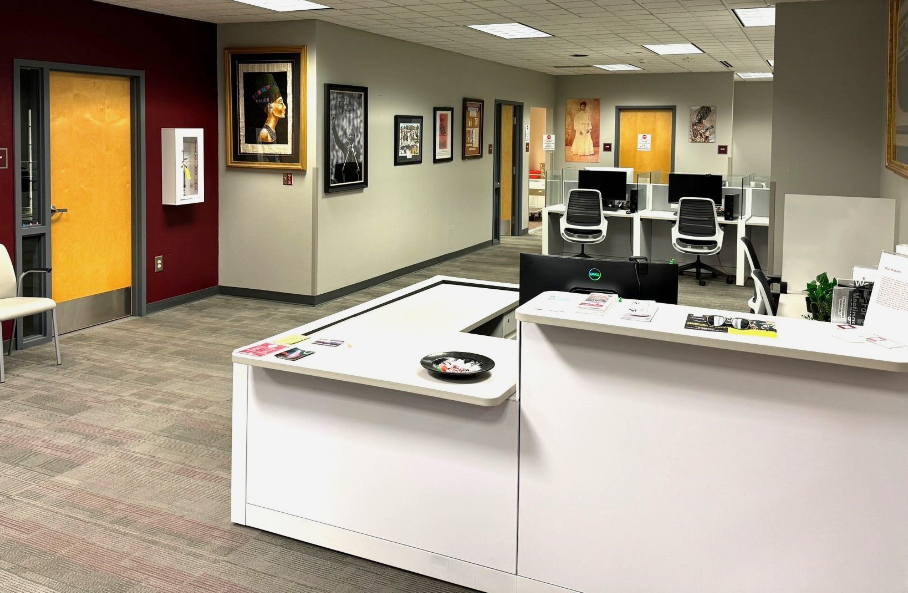 The image shows the interior of an office space. In the foreground, there is a white reception desk with various pamphlets and a bowl with candies. Behind the desk, a computer with a monitor is visible. The flooring is carpeted with a pattern of gray tiles.  To the left, there's a door with light wood paneling and gray trim. Beside the door, a white fire extinguisher case is mounted on a maroon-colored wall. A beige chair is positioned against this wall near the door.  The background reveals an open office layout with several white desks set against neutral-toned walls. Each desk is equipped with a white office chair and computer monitor. The ceiling is made up of white tiles with embedded lighting fixtures.  The walls are adorned with framed artwork and posters. Notable pieces include a portrait of a figure in traditional attire, various black-and-white photographs, and colorful prints. There are also additional office spaces or rooms visible through open doorways in the background.