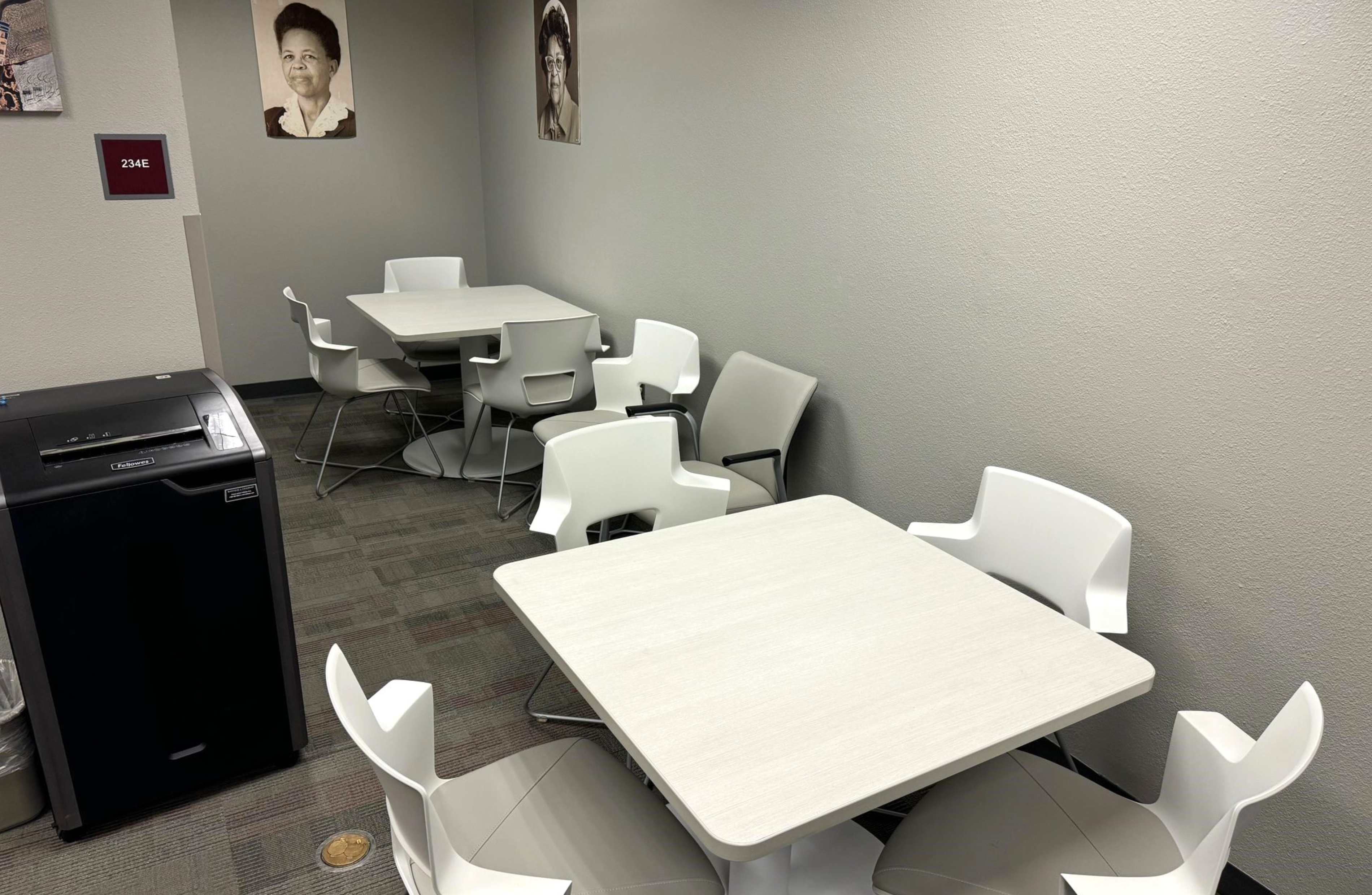 The image portrays a small, furnished room with light grey walls and a grey, carpeted floor. There are two square tables, each surrounded by four chairs. The chairs are a mix of white plastic ones with armrests and beige cushioned ones with black armrests. On one side of the room, there is a black paper shredder with a silver trash can beside it. Mounted on the left wall is a red sign with white text. On the walls are two large, black-and-white photographic portraits of individuals.