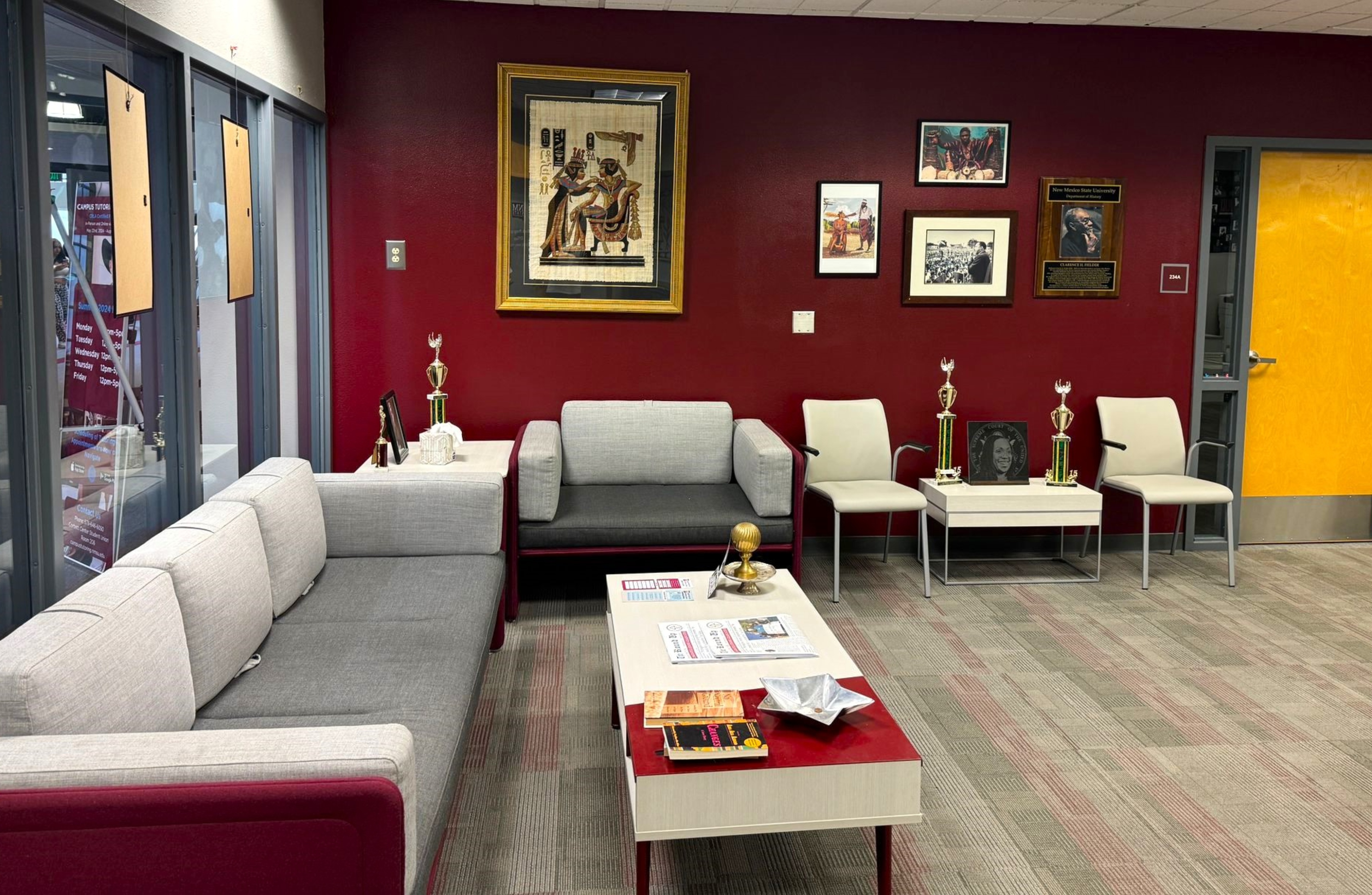 The image depicts a modern waiting area with a mix of comfortable seating, a coffee table, and various decorative and accolade items. The room has a warm and inviting atmosphere with a bold contrast of colors and textures.  Overview:  Seating Area: The seating includes light grey sofas with maroon sides and accents and light grey armchairs. Coffee Table: A rectangular coffee table with a white top and red accents holds various items including books and pamphlets. Decorations and Accolades: The wall behind the seating arrangement is painted maroon and features a collection of framed pictures, plaques, and artworks, along with several trophies placed on side tables. Other Features: The room has large windows on the left side giving a glimpse into another area with informational posters. A yellow door is positioned on the right side of the back wall. Details:  Light Grey Sofas: Positioned against the left and middle parts of the room. Coffee Table Contents: On the table are several pamphlets and a few books. Among them is a book titled "Existentialism and Human Emotions." Framed Pictures and Artwork: The wall displays various framed pictures including historical photos and a large framed artwork depicted in a gold frame. Side Tables: There are side tables next to the sofas and armchairs holding trophies and other small decor items. Trophies: The trophies are golden and silver in color and positioned on the side tables beside each type of seating.