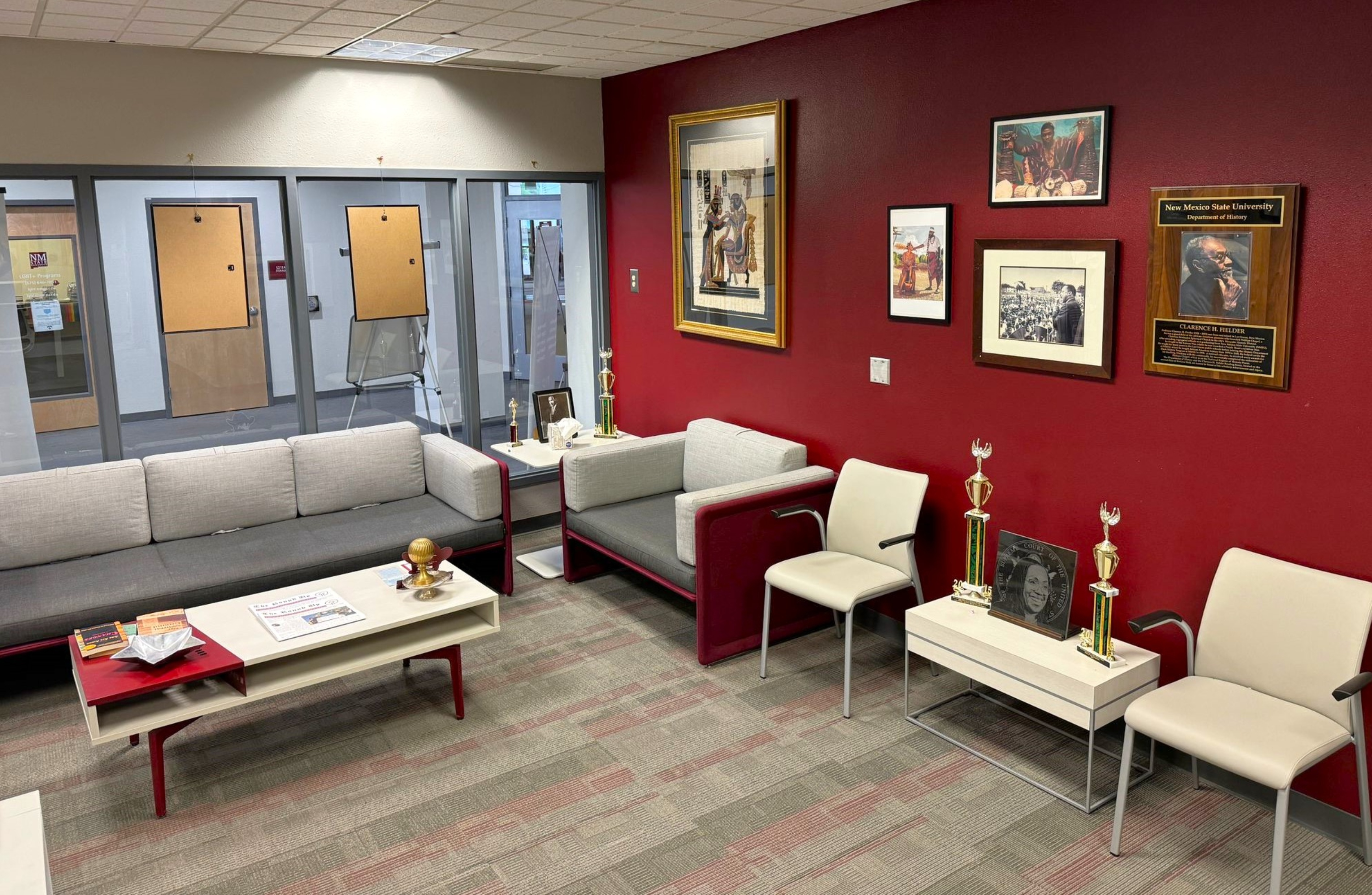 The image depicts a waiting area inside an office. The room features a mix of grey and red furniture, including a grey sectional sofa with red armrests and two white side tables. One side table has a trophy and some papers on it, while the other holds a small bronze bust, an award plaque, and another trophy. In front of the sofa is a rectangular white coffee table with a shelf underneath, displaying various magazines and a small golden globe ornament.  The back wall of the room is painted red and decorated with a number of framed photographs and a plaque. This wall stands in contrast with the other walls, which are light grey. The floor is carpeted with a pattern of grey and red rectangular tiles. Opposite the red wall are three glass office doors with beige-colored bulletin boards attached to them. Beyond these doors is another portion of the office space.