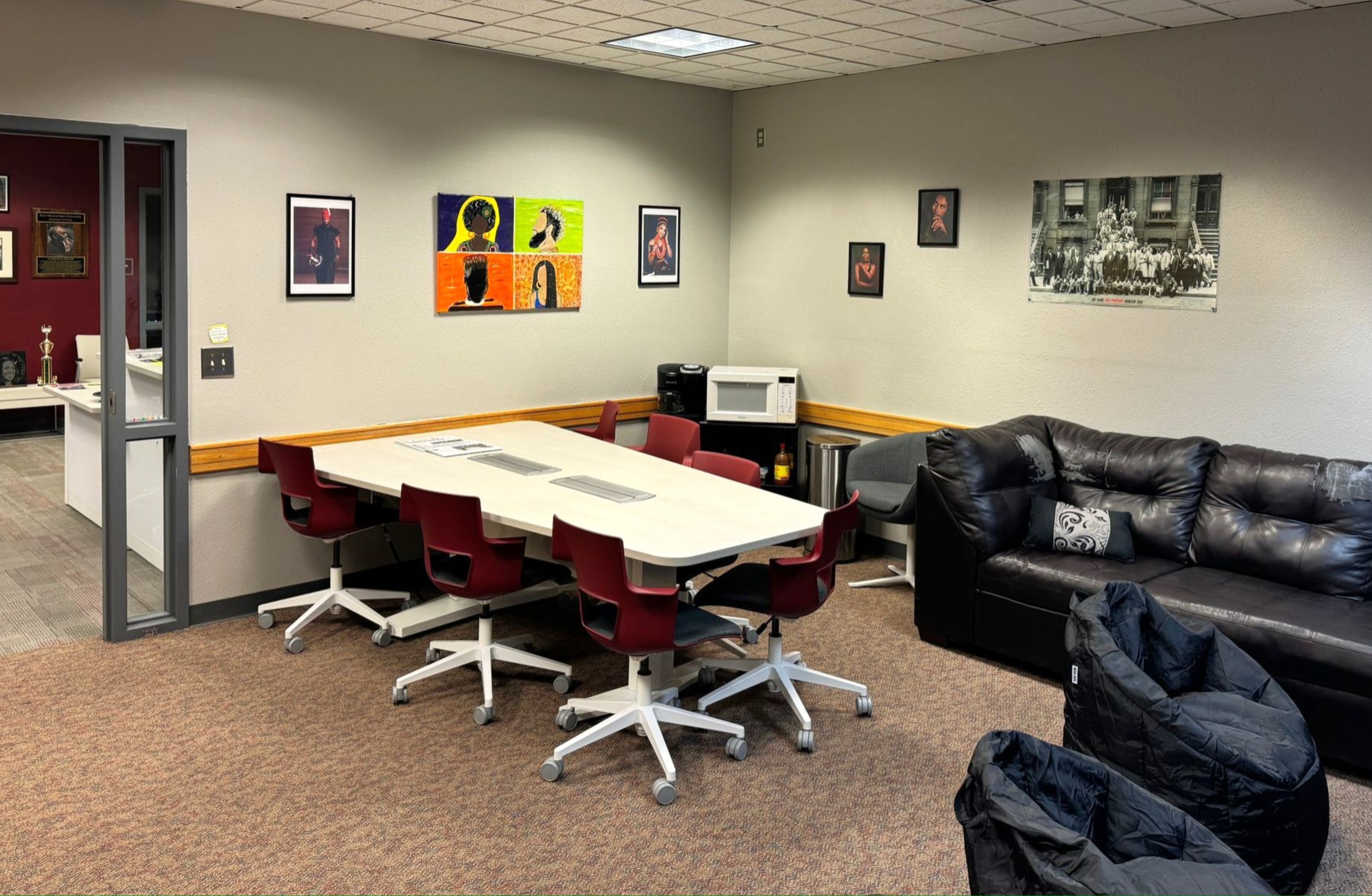 The image showcases a well-organized, multipurpose room with a combination of a conference area and a lounge. The room has beige carpeted flooring and light grey walls. Ceiling tiles form a grid on the otherwise plain ceiling, with fluorescent lighting providing illumination.  In the left portion of the room, a sectional, white, rectangular conference table surrounded by six red swivel chairs with white bases is present. Adjacent to this, the right wall is adorned with two framed photographs and a vibrant, multi-colored painting depicting six different stylized profiles of heads and faces. The far right of the wall hosts more art, including a monochromatic old photograph of a large group of people in front of a building, and two smaller framed pictures featuring individuals.  Adjacent to the conference area, there's a white microwave, a black coffee machine, and a small counter. In this corner, a grey armchair sits next to a white trash bin.  Along the right wall, a large black leather couch with decorative cushions faces a pair of black bean bag chairs indicating a lounging area.  In the background, there's an office or possibly another room, visible through an open glass door, which contains more desks, chairs, and a trophy.